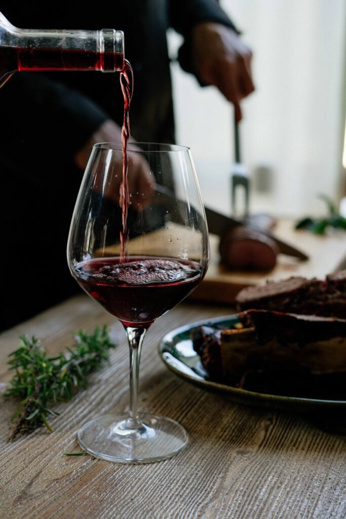 wine being poured form a glass with a plate of meat in the foreground, and somone cutting something in the background the back ground is blurrred