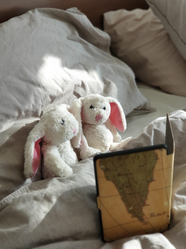 2 stuffed animals (white rabbits) on a bed with grey sheets reading a book 