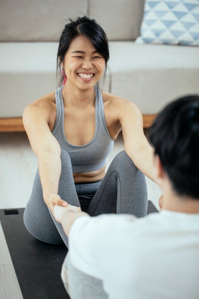 Cheerful young Asian woman in sportswear smiling and holding hand of anonymous boyfriend while practicing yoga together on mat in living room