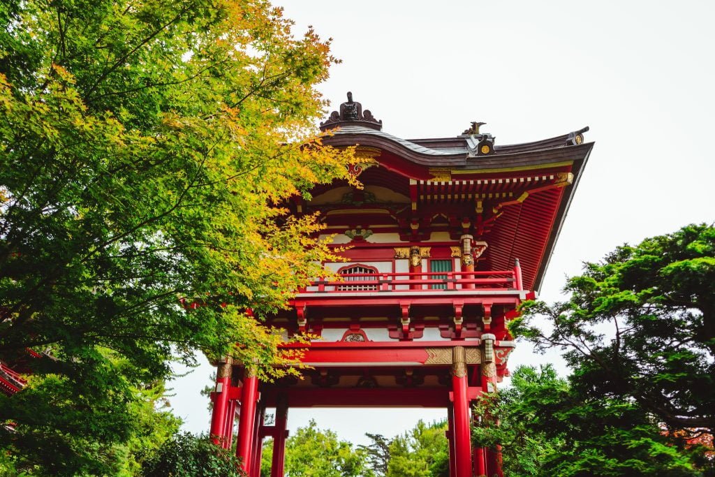 Low angle exterior of traditional aged red Asian temple surrounded by lush green trees in Japanese Tea Garden located in San Francisco on sunny day